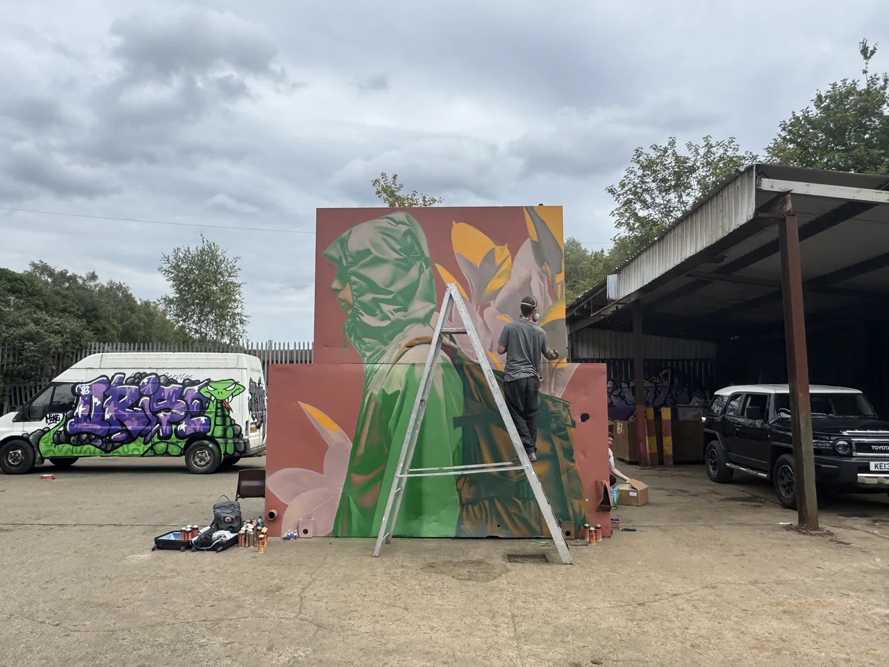 A tall outdoor panel painted with angular green and orange shapes beside a ladder.