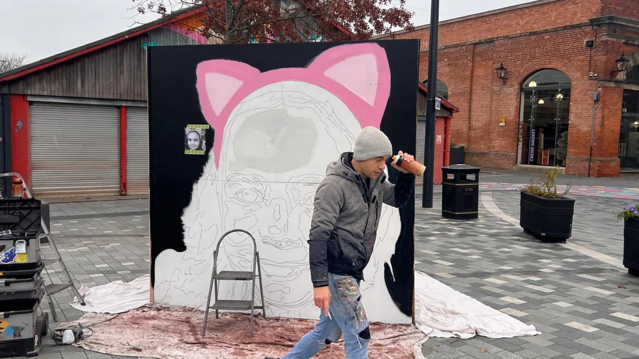 A painter holding a spray can in front of an outlined portrait with pink cat ears in Lyceum Square.