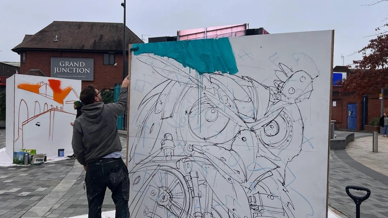 A painter adding turquoise paint above an outlined owl on a freestanding panel in Lyceum Square.