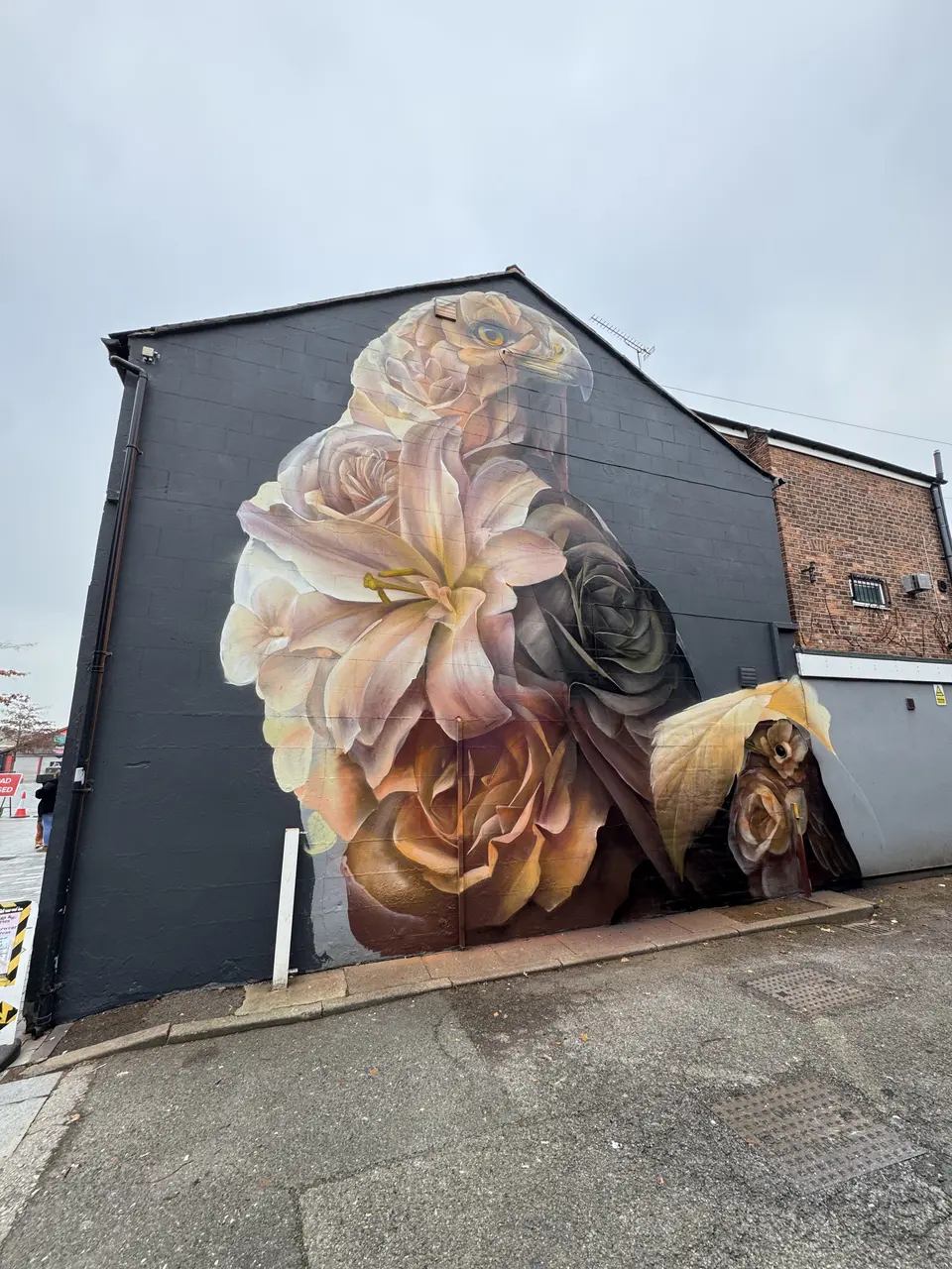 Large gable-end mural on a dark wall: a bird of prey formed from cream lilies and roses, with a small owl among golden leaves at its base.