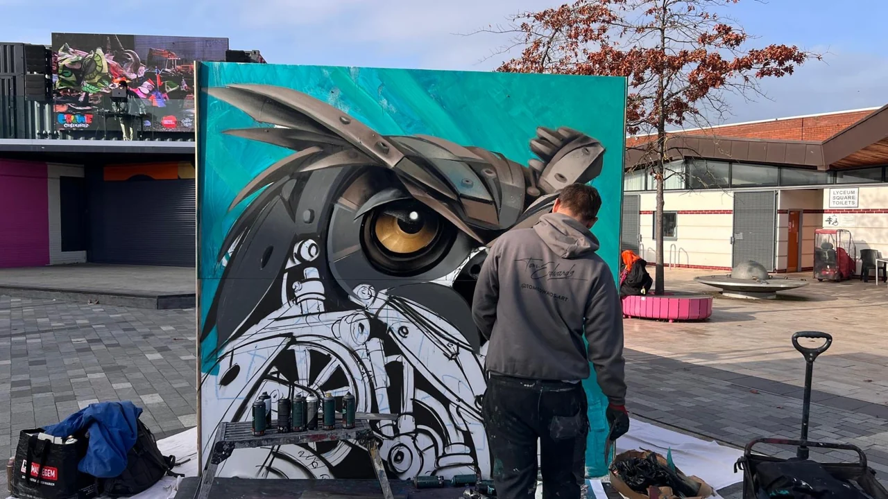 A painter working beside a detailed owl painted on a turquoise panel in Lyceum Square.