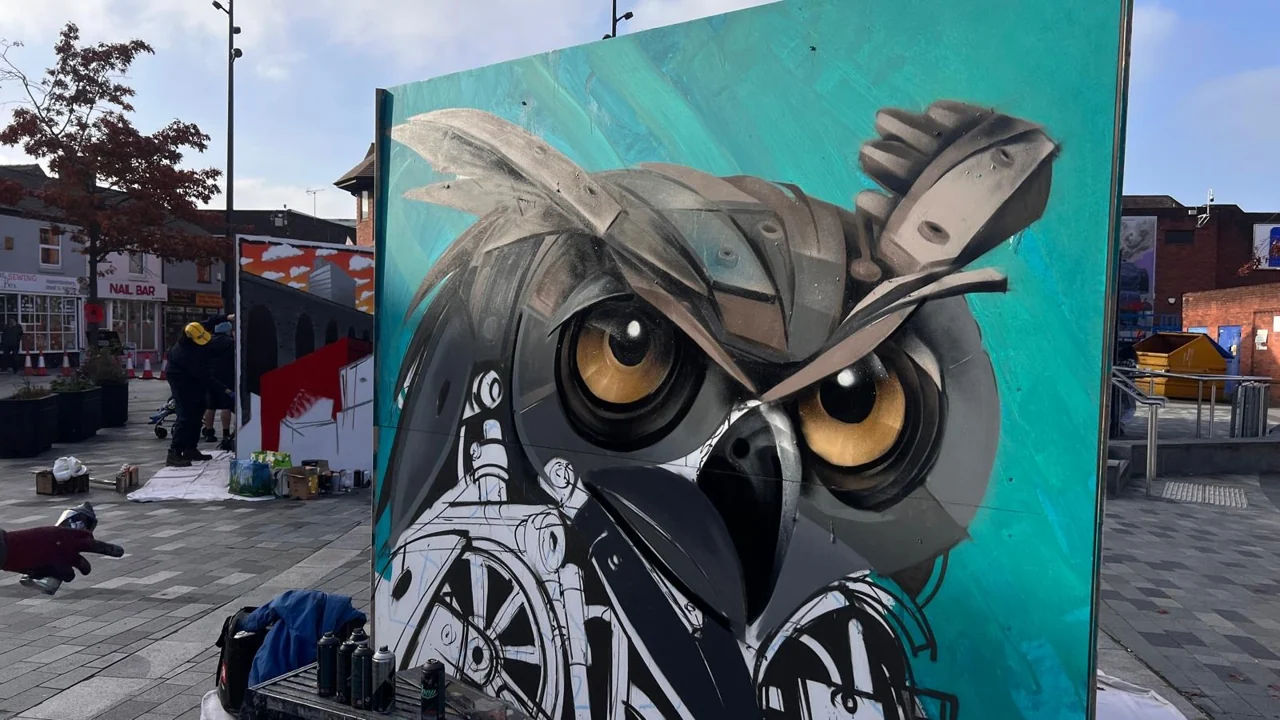 An owl painted on a turquoise freestanding panel in Lyceum Square, with a painter working beside it.