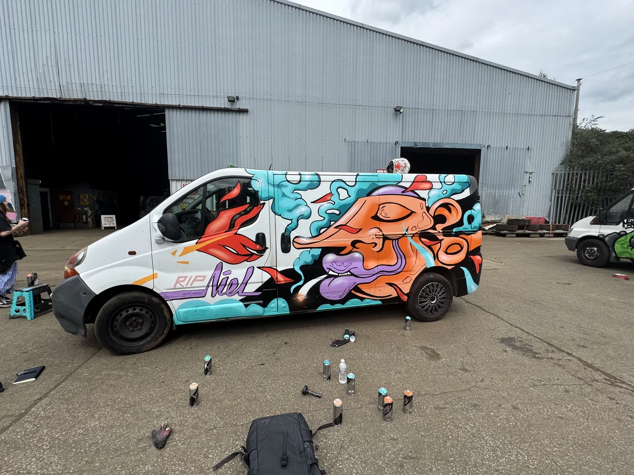 A white van covered in orange and turquoise graffiti with spray cans nearby.