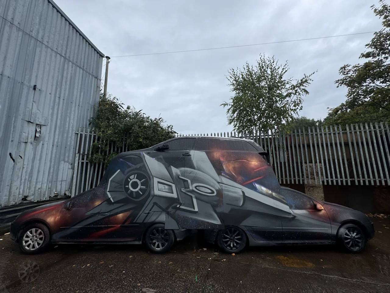 A dark car painted with a space-themed design beside corrugated buildings.