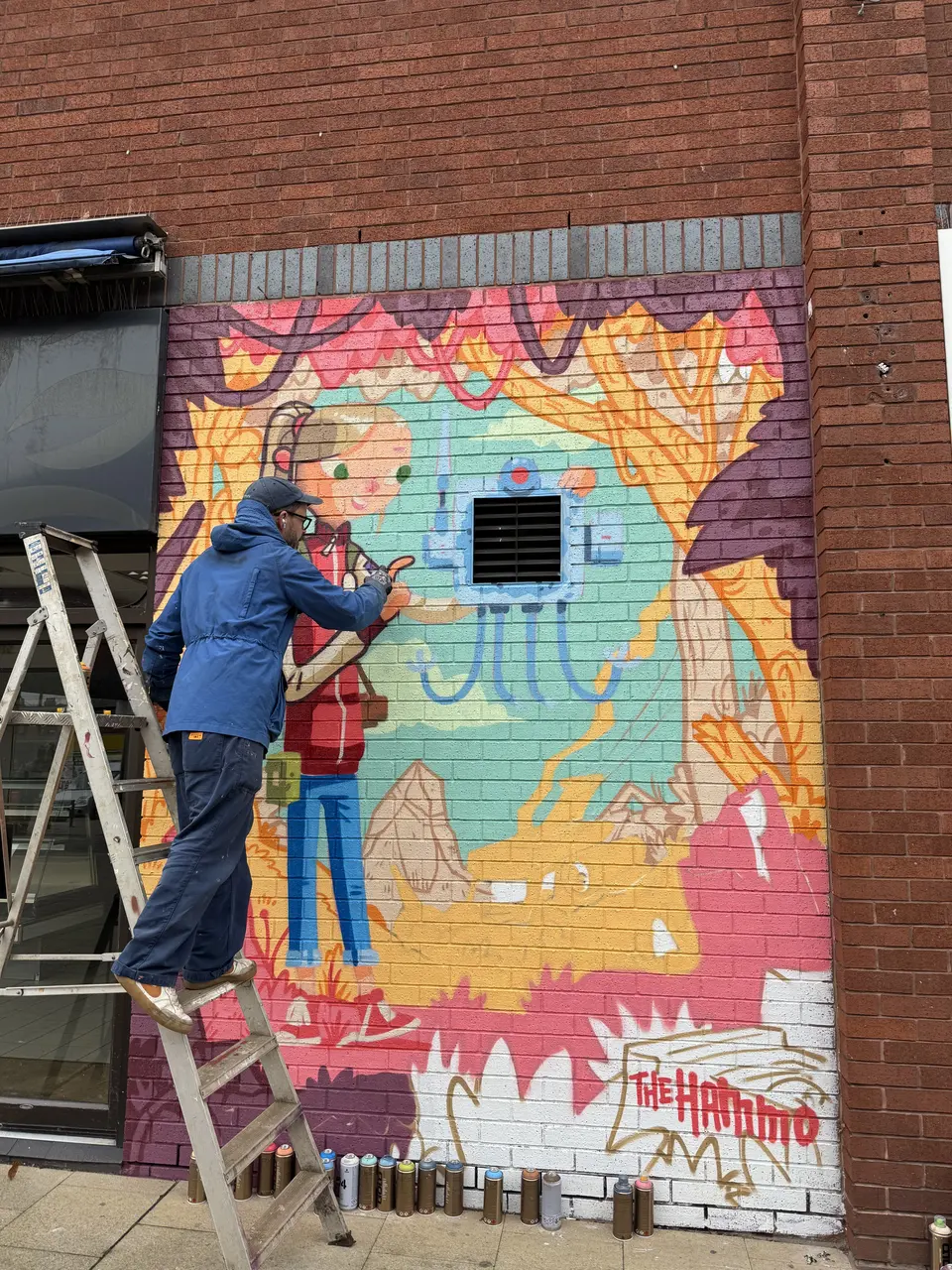 An artist on a stepladder paints a colourful brick-wall mural in pink, teal and yellow. Spray cans line the pavement; the wall is signed 'The Hammo'.