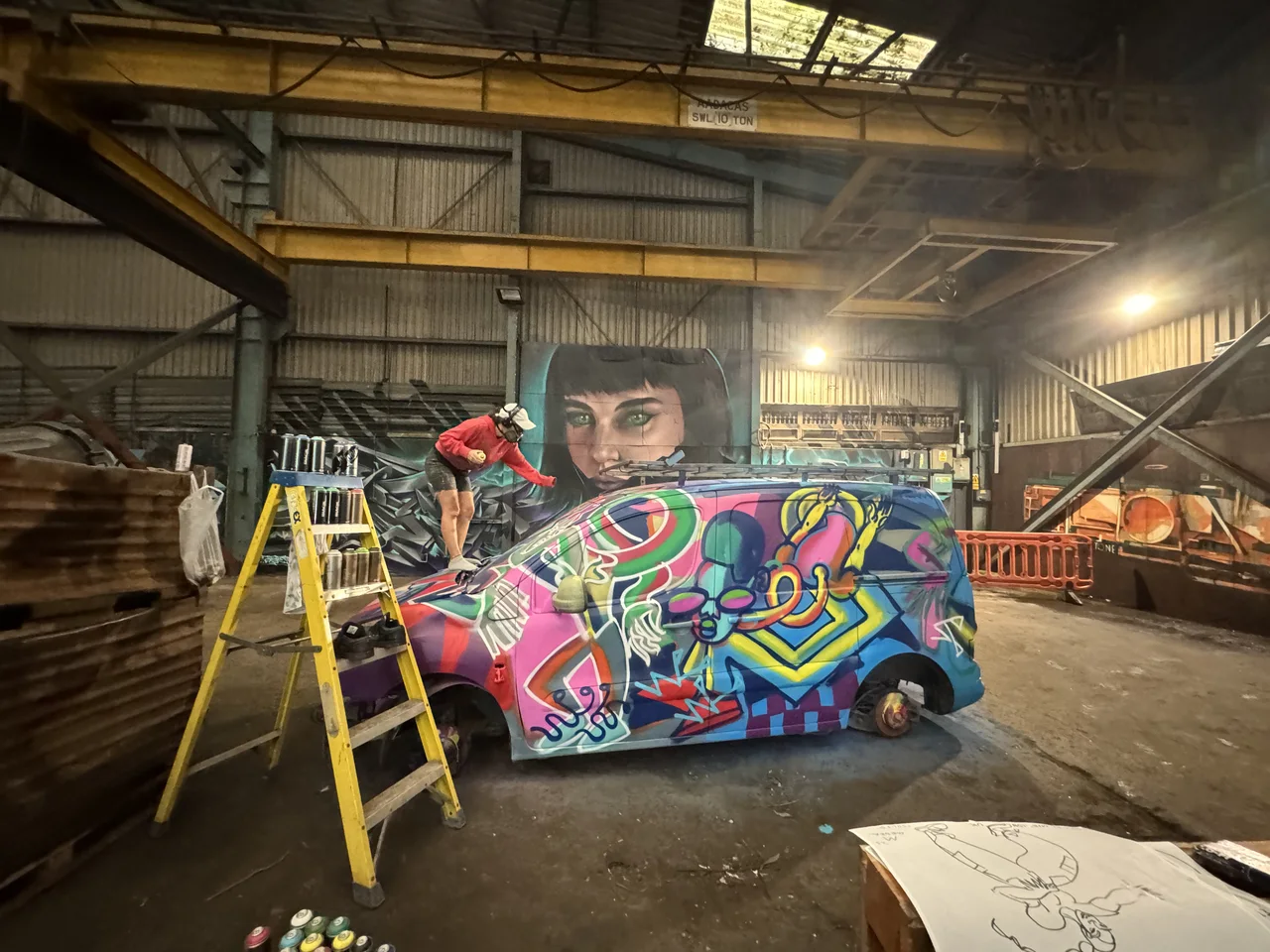 A colourful painted vehicle beside a stepladder inside the scrapyard workshop.