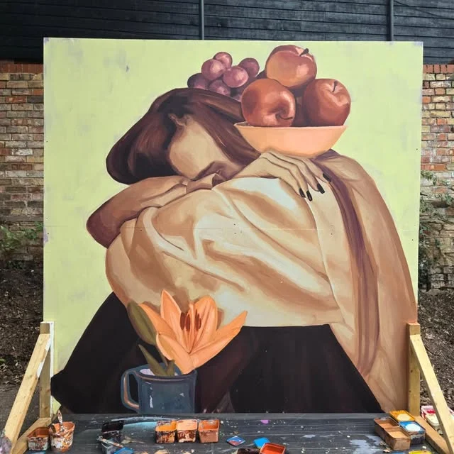 Doppel portfolio mural: two embracing figures beneath a bowl of fruit, with an orange lily, on a pale green wall.