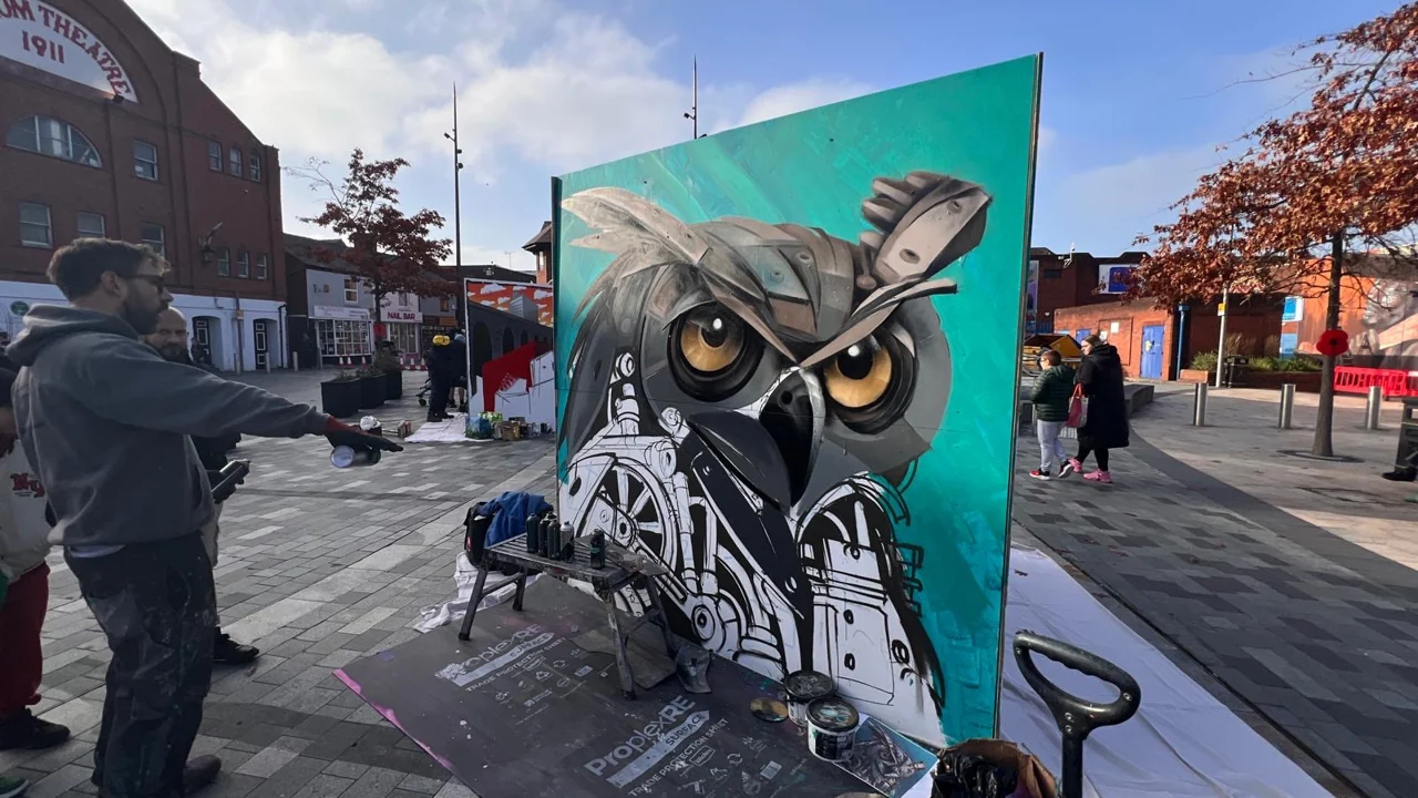A wide view of an owl painting on a panel in Lyceum Square, with people and painting supplies nearby.