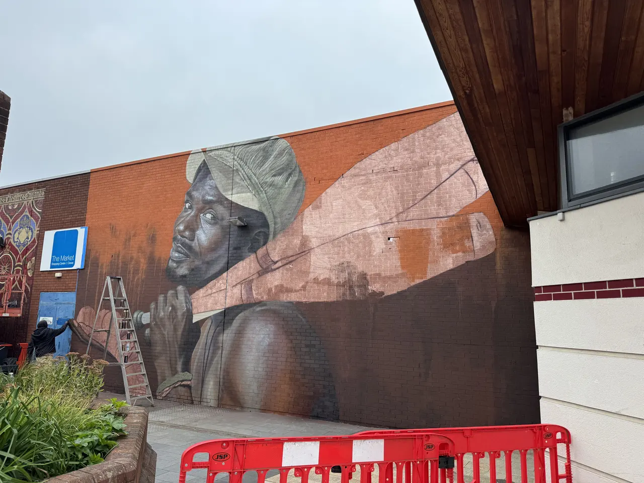 Work in progress: a large photorealistic portrait of a man in a cap carrying a long wooden beam, painted across a rust-red brick wall behind safety barriers. A ladder stands at the wall.